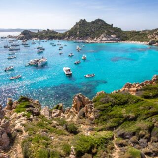 The crystal clear waters of Sardinia with a rugges coast filled with rocks and grass. You can also see sail boats floating in the water off the coast if you rent a car in Sardinia.