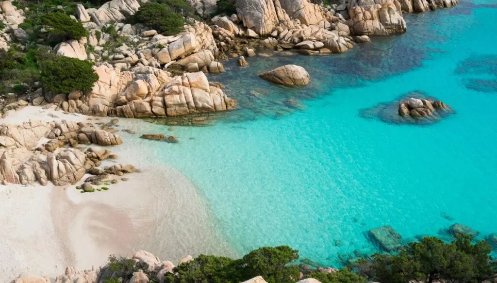 Aerial view of a pristine white sand beach with clear blue water in sardinia. It is surrounded by light rocks and lush greenery. 