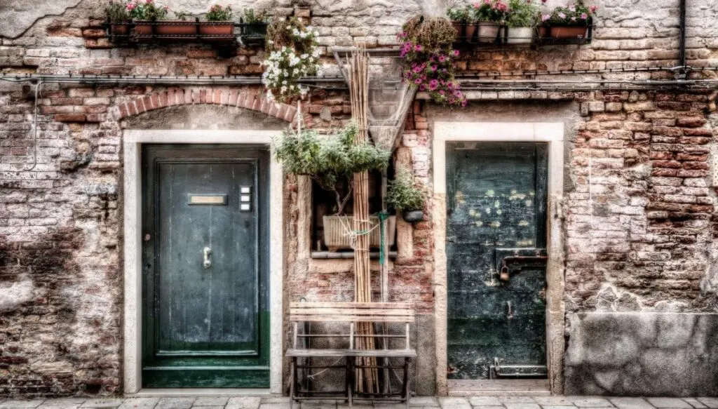 A view of two old, black doors in an old brick building in the Cannaregio neighborhood of Venice. They are surrounded by plants with flowers. 