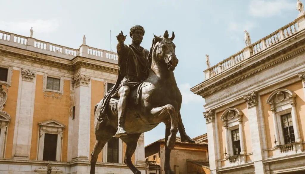 View of the bronze Marcus Aurelius statue where he is riding a horse in Campidoglio Square. He is surrounded by yellow buildings on top of Capitoline Hill.