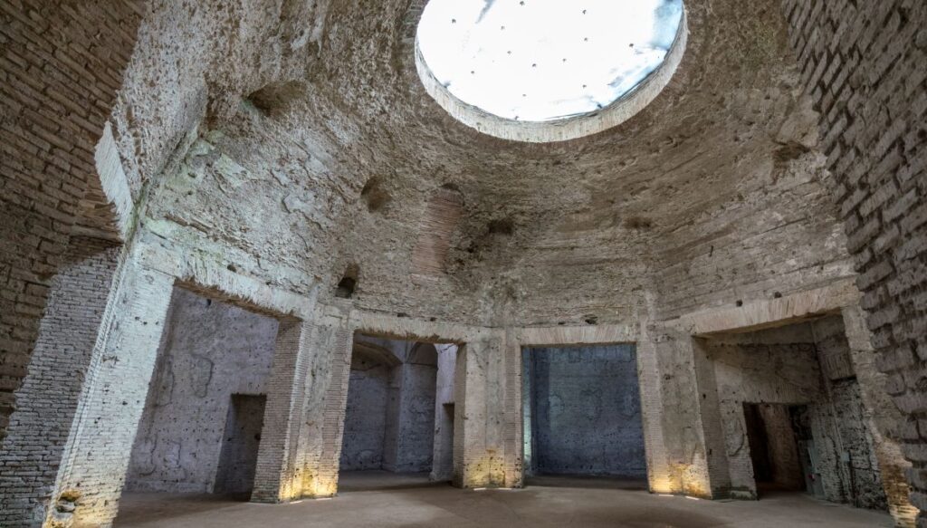 A view of the interior of the dome that forms the ruins of Domus Aurea. It is a circular, white brick building with a circular hole in the roof and four square doorways along the floor of the building