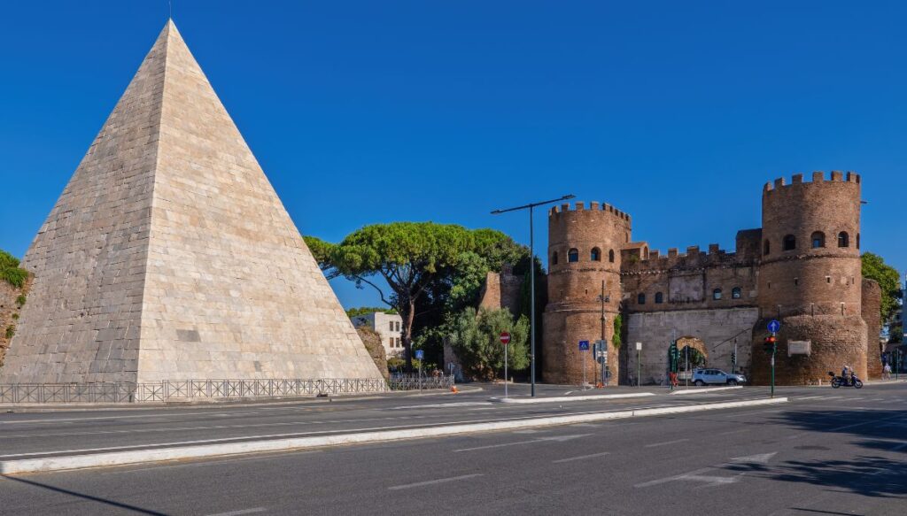 A giant white pyramid sits next to the San Paolo gate which features two circular towers made of brick with a connecting arch. 