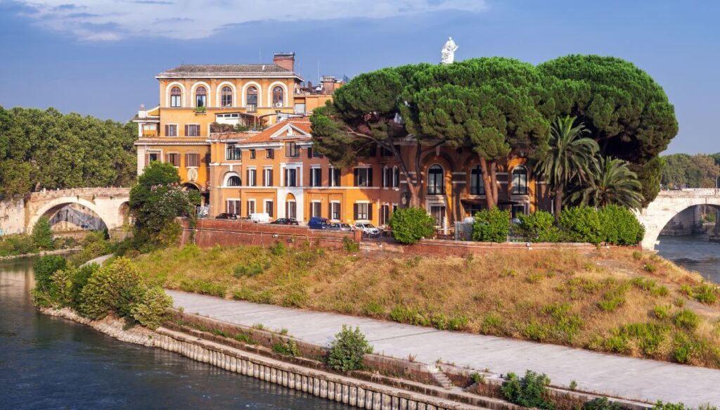 A view of a yellow mansion sitting on Tiber Island in Rome. Arched bridges connect the island to the mainland in either direction.
