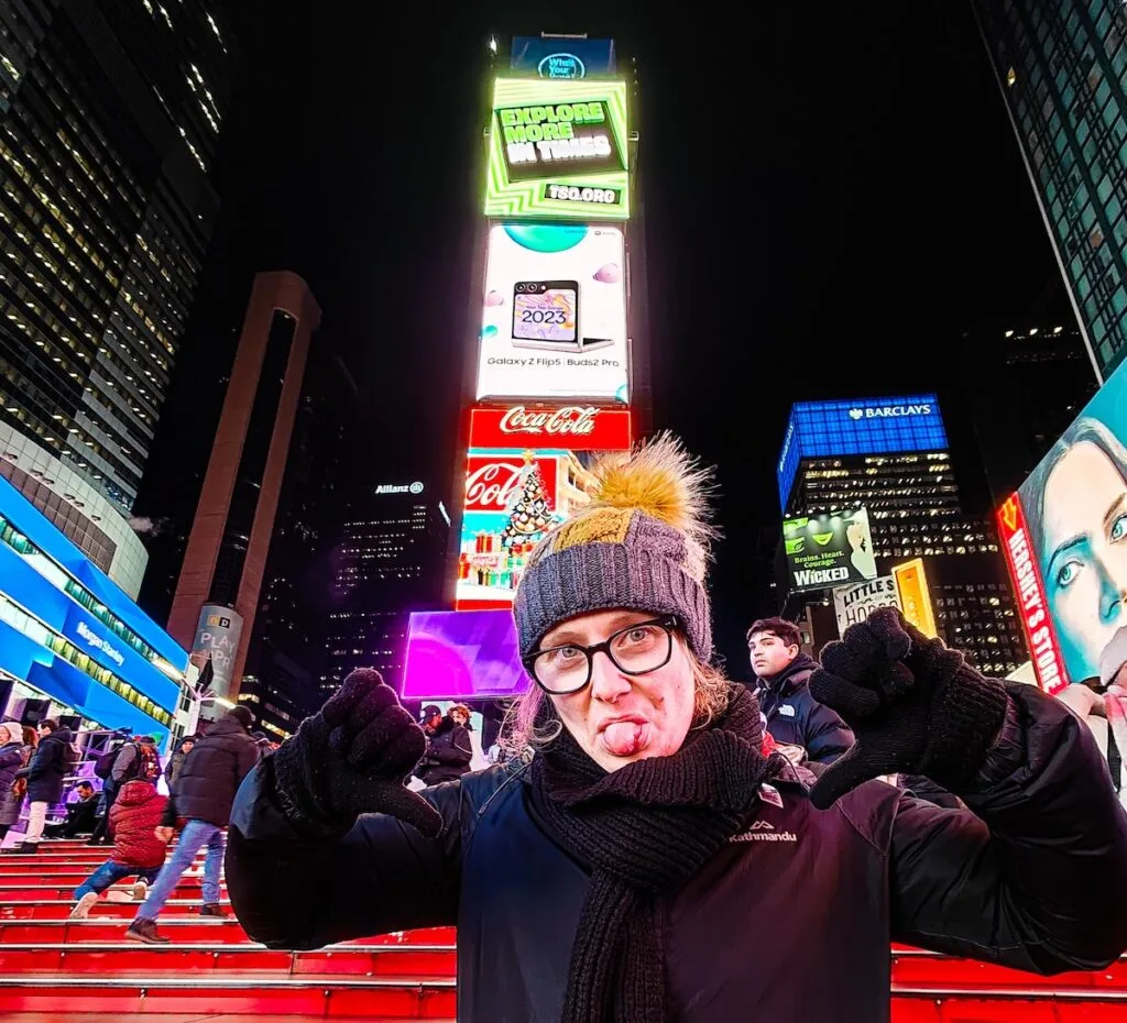 Me standing on the red steps in Times square with my arms bent and near my ears and my toungue sticking out. I am standing in front of a neon sign and I have a gray and yellow hat with a black jacket.