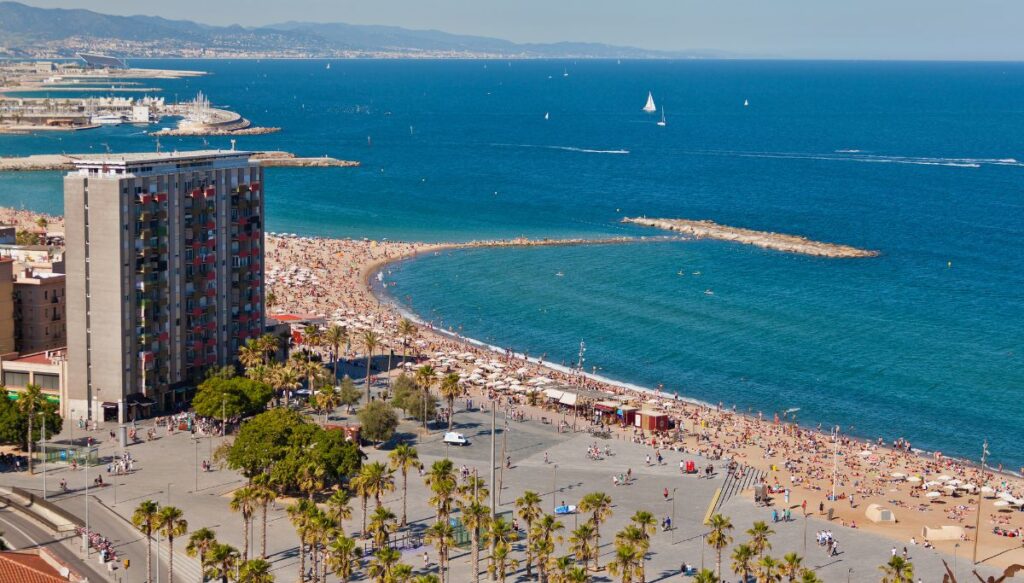 Aerial view of Barceloneta Beach in Barcelona. You can see the bright blue water and people sitting on the sand. the beach is surrounded by tall skyscrapers.  