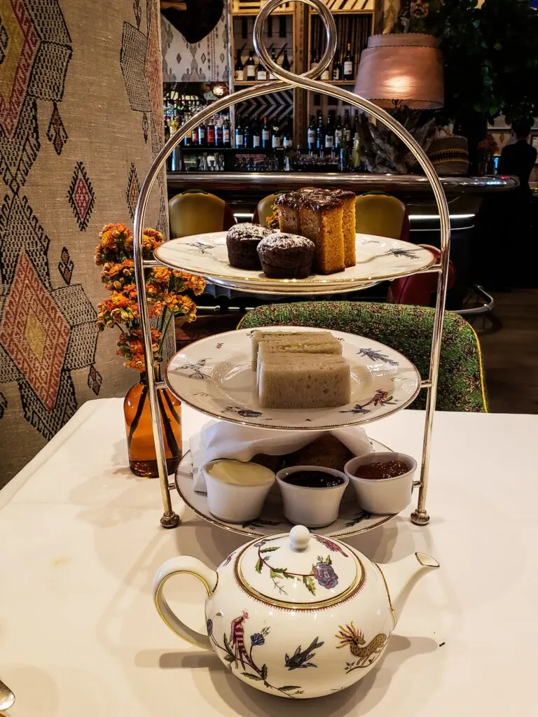 A three-tiered afternoon tea plate with a metal handle and white places. The top plate has cakes, the second plate has tea sandwiched, and the bottom plate has scones and jam. A floral, white teapot sits infron on a white tablecloth and theire is a vase with flowers on the left. 