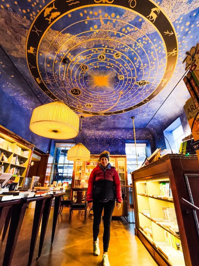 Me standing in a winter coat and black leggings with the constellation studded ceiling of Albertine bookstore above me. The ceiling is bright blue and there are stars everywhere. I am surrounded by circular lamps hanging from the ceiling and shelves/tables of books.