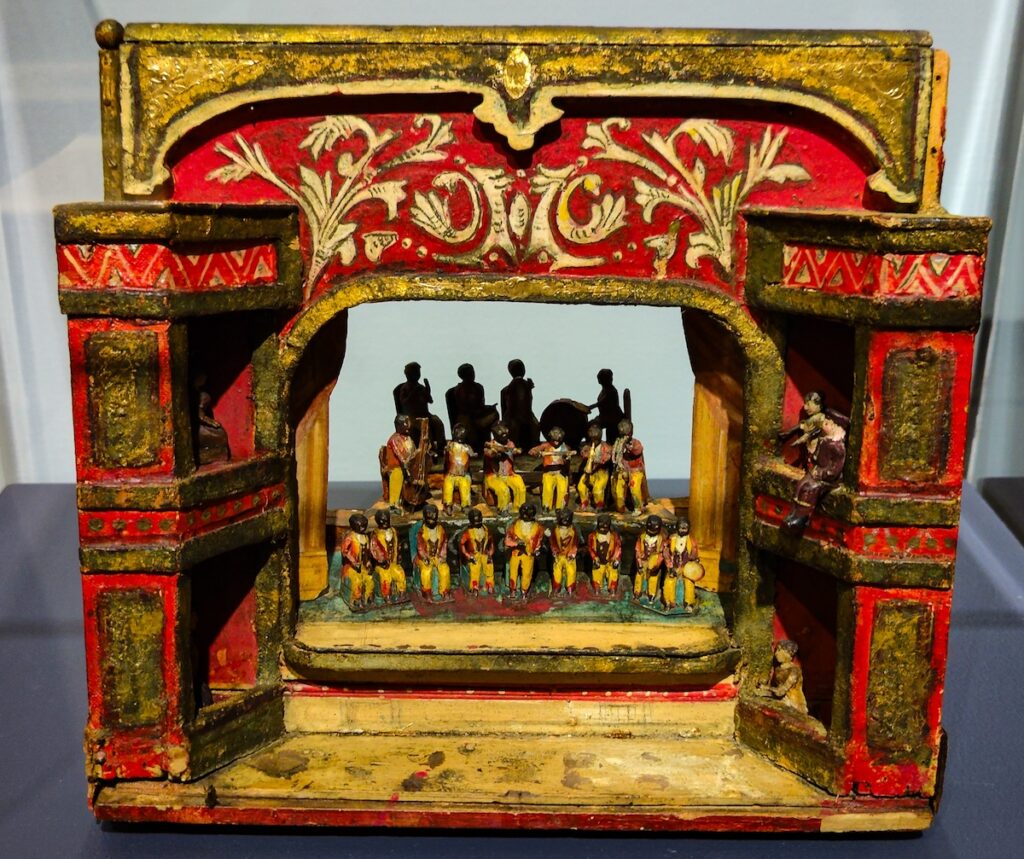 View of a miniature red stage with miniature poeple all lined up in the center with red curtains are the gold stage. This American piece of art is kept behind glass at the American Folk Art Museum. 