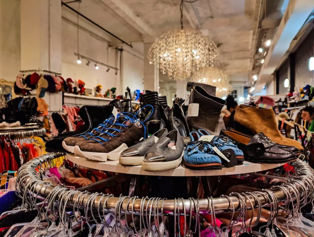 An assortment of shoes sitting on a circuliar rack with hangins filled with clothes. It's a large white store with crystal chandeliers in the background. 