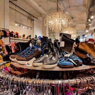 An assortment of shoes sitting on a circuliar rack with hangins filled with clothes. It's a large white store with crystal chandeliers in the background.