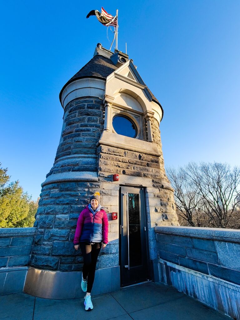 Me standing in a pink/blue winter coats and turquoise sneakers,a nd black leggings in front of Belvedere Castle. I am standing againsta. stone tower with one leg bent and there is a US glad waving from the top of the tower on a clear day. 