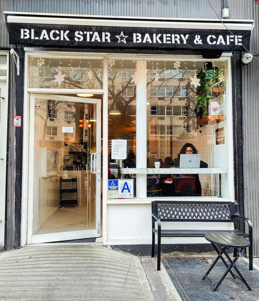 The exterior of Black Star Bakery. It has a black awning, a white bnch out front, and lots of windows. You can see a women working on her laptop through the window, on the right. 