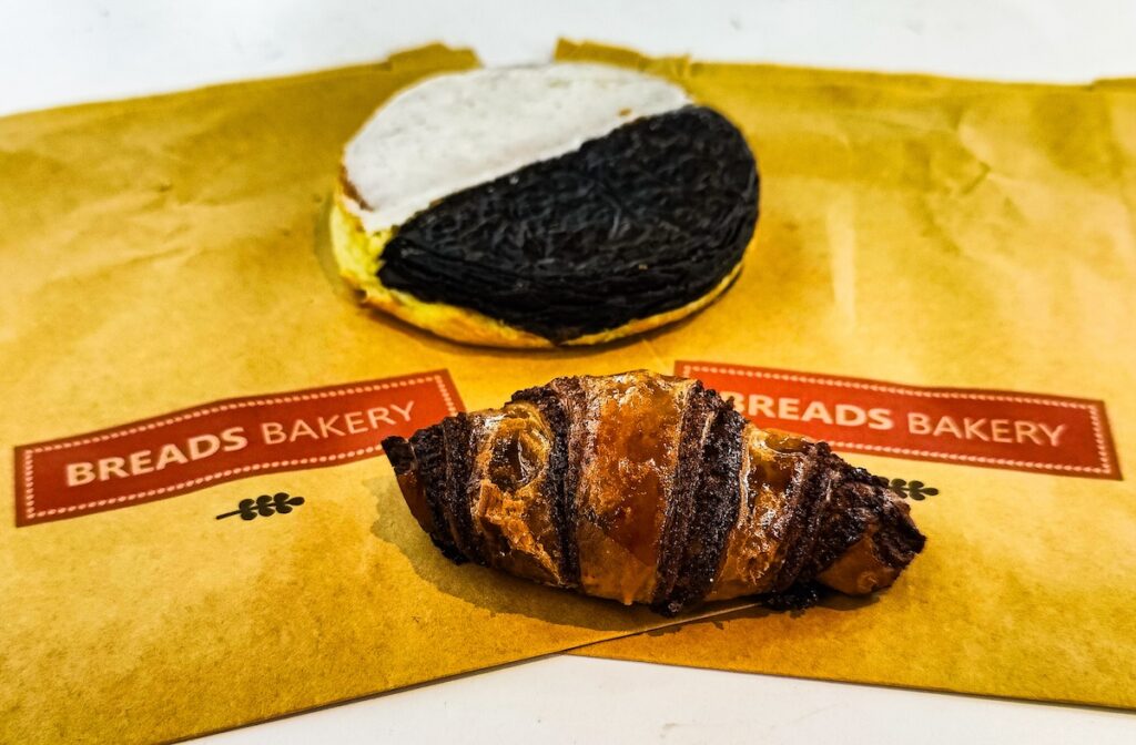 Picture of a black and white cookie on top and a piece of chocolate rugelach beneath at one of the best dessert places in NYC. They both sit on a brown bag with the Bread Bakery logo on top. 