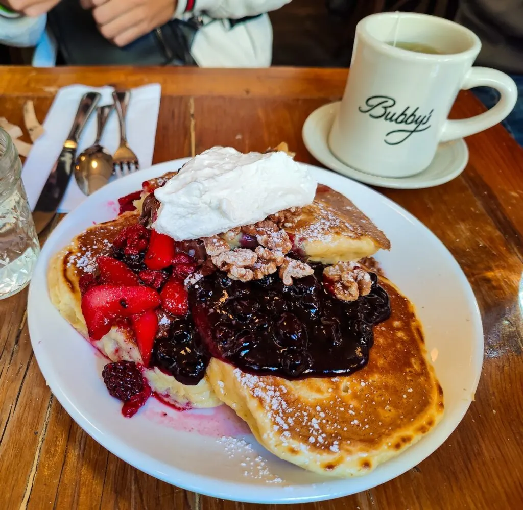 Plate of three giant pancakes served with whippers cream and berry compote on a wooden table at Bubby's. You can see a white mug with the Bubby's logo on it in the background as well as some silverware on a napkin. 