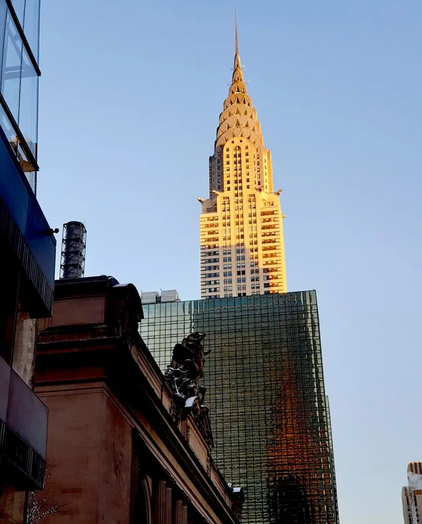 View of the Chrysler Building all lit up at sunset. It is a triangular skyscraper with triangles around the toip. It is surrounded by buildings and this view is from the street below on a clear day in NYC. 