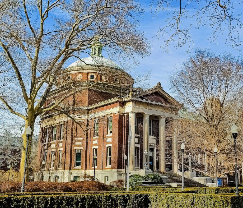Circular brick building with four white columns out front. There it a domed roof and then a triangle above the columns. The historic building is surrounded by trees and bushes.