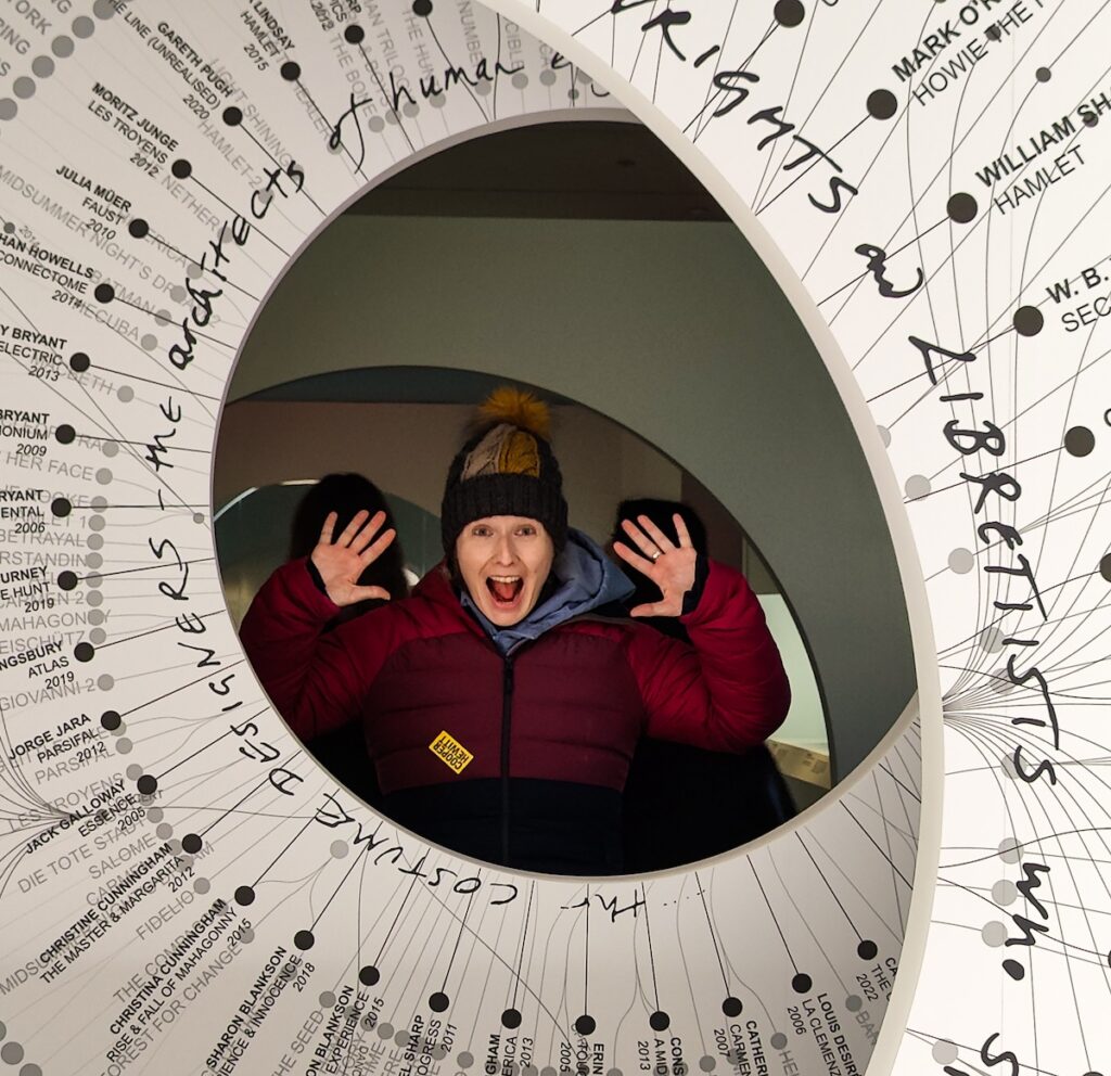 Me enjoying a temporary exhibit at the Cooper Hewitt. I am surrounded by a circle of white that has black writing on it. I am in a purple and blue coat and have a winter hat on with my elbows bent and my hands up by my ears. I have a surprised look on my face. 