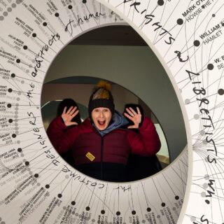Me enjoying a temporary exhibit at the Cooper Hewitt. I am surrounded by a circle of white that has black writing on it. I am in a purple and blue coat and have a winter hat on with my elbows bent and my hands up by my ears. I have a surprised look on my face.