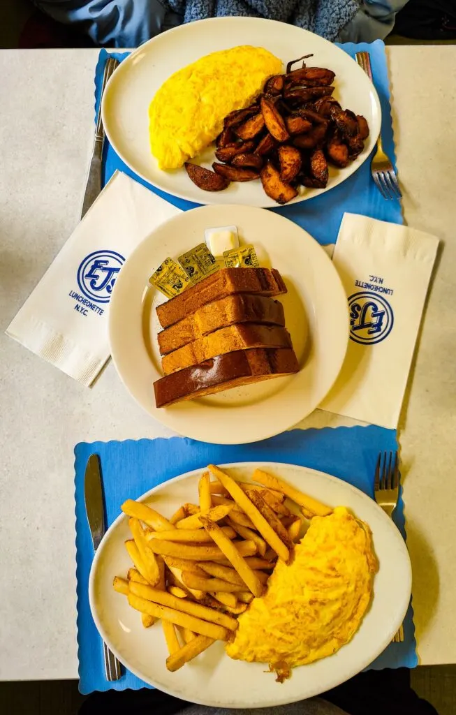 Aerial view of a white table with two omelets. One has homefries on the plate and the other has french fries. They sit on two blue placemats and there is a plate with four pieces of Challah on it in the middle of the table. That Challah has two EJ napkins on either side. 