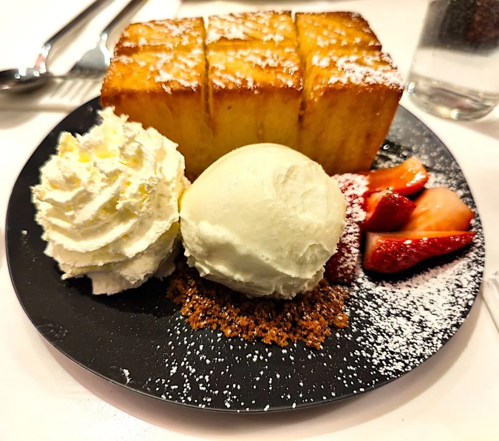 View of the Golden Toast from Spot Dessert Bar. it is honey coated toast on a black plate with a scoop of ice cream, strawberries, and whipped cream. It is on a white table and surrounded by silverware. 