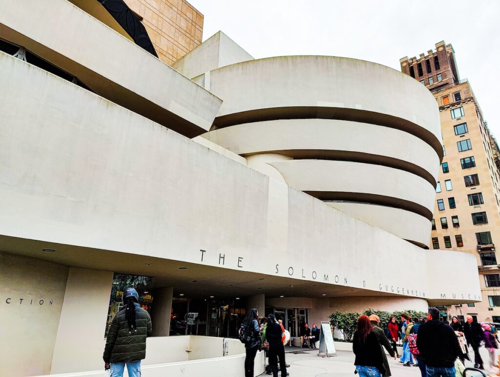 A view of the modern, white exterior of the Guggenheim. It has lots of clean lines and a curved part as people walk by up and down Fifth Avenue. 