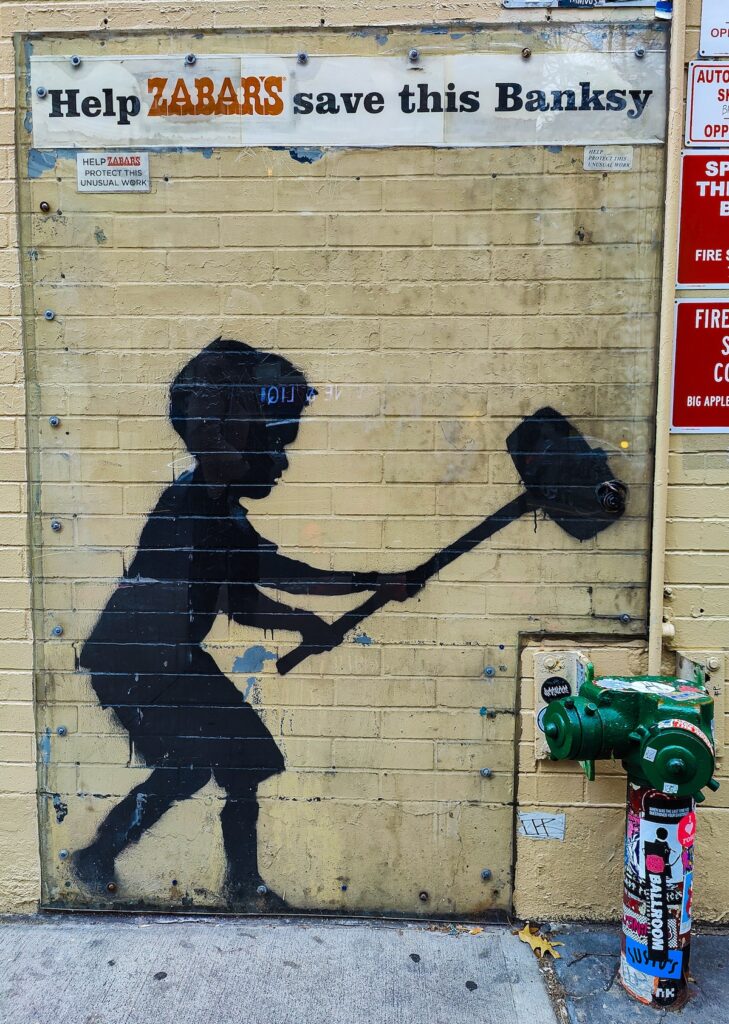 You see the black silhouette of a little boy holding a hammer and getting ready to hit a green fixture on the building. It is by Banksy and on the wall of a yellow brick building and is protected by a plastic cover.