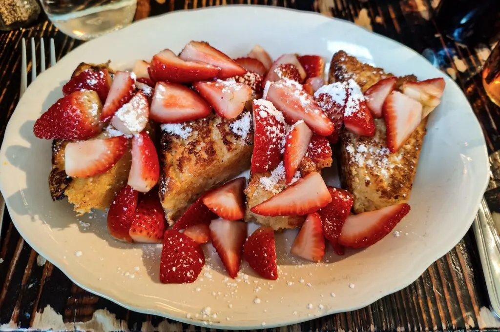 Four giant pieces of French Toast covered in powdered sugar and strawberries on a white plate. It sits on a wooden table with a silver knife and fork on either side.