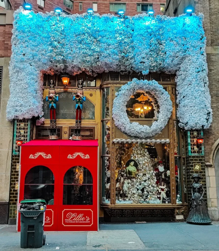 The exterior of Lillie's Victorian Establishment has tons of white flowers all along the top and a white wreath just below it. A red, box-like awning stand in front of the door. Two toy soldiers sit above the door and a white Christmas tree sits in the window.