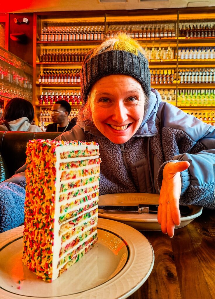 Me sitting at a table with a 24-layer funfetti cake in front of my. I am wearing a blue fleece and a gray and yellow hat with a wall of bottles behind me at one of the best dessert places in NYC.