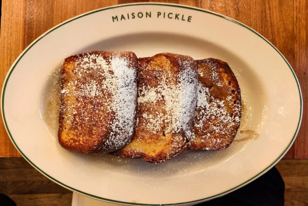 Aerial view of a white , round plate at Maison pickle with three slice of Challah French toast with syrup and powdered sugar. They sit on a wooden table at one of the best brunch places NYC has to offe.r