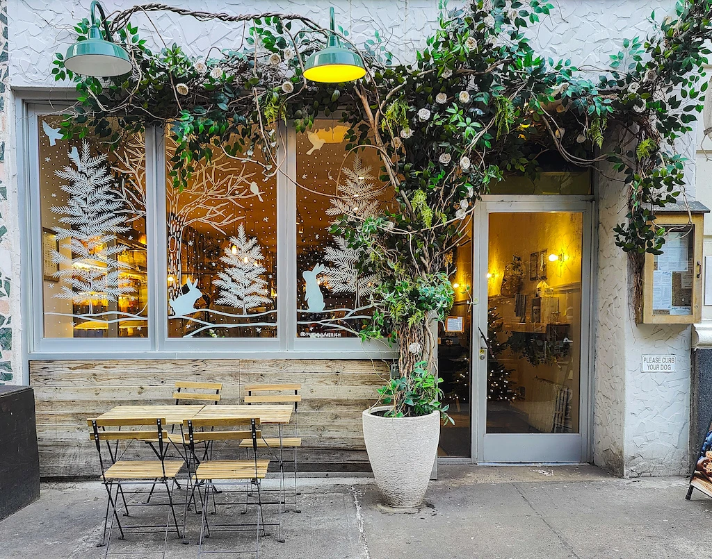 The white exterior of maman is adorned with green leaves and white trees on the windows. A wooden table and chair also sits outside the front door. 