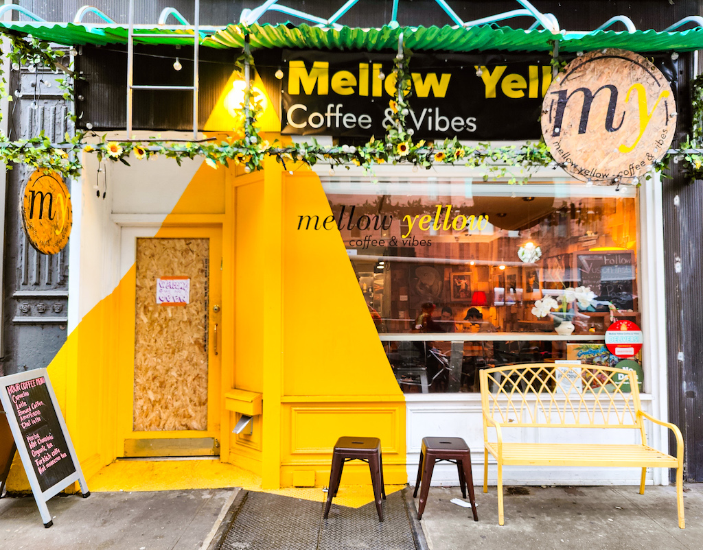 A view of the exterior of Mellwo Yellow. There is a yellow triangle over the door, which is boarded with wood. There is also a window accented by white and a yellow bench out front. 
