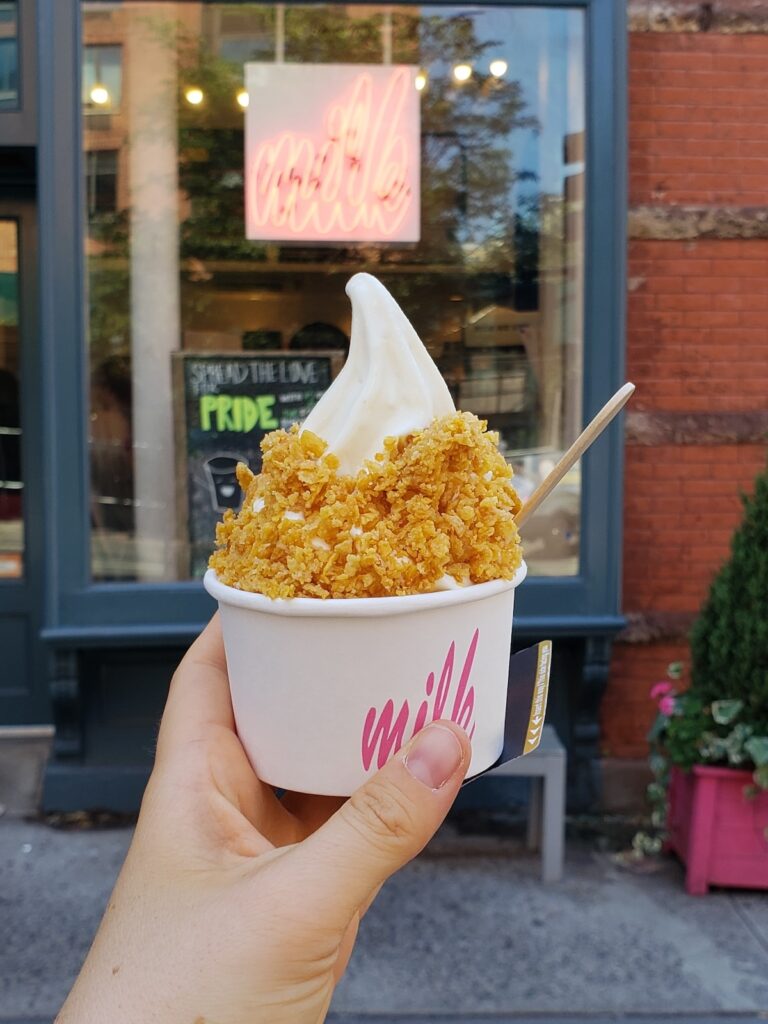 Me holding a ceral and milk soft ice cream in a cup in front of the sign for Milk Bar in NYC. 