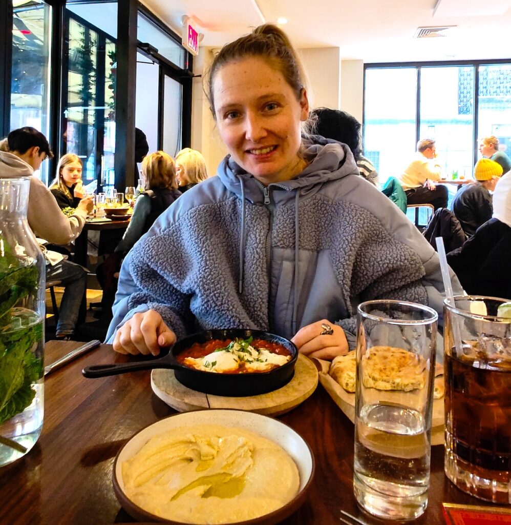 Me sitting behind a table in a blue fleece with a plate of Shakshuka and a bowl of humus with olive oil and water with mint on a table in a light and airy dining room.