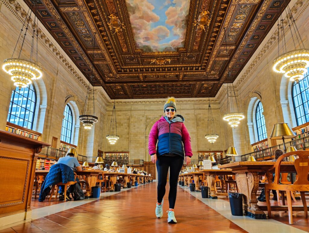 Me enjoying the Rose Main Reading Room in the New York Public Library. I have a winter jacket, a hat, and green sunglasses on and am walking towards the camera. I am surrounded by wooden work desks and there is a ceiling mural with ornate wood trim.