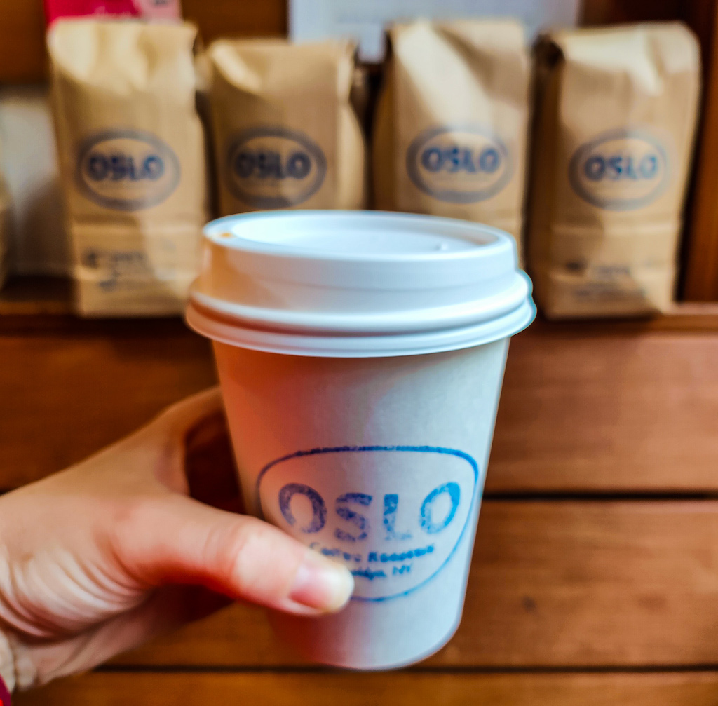 Me holding a white to-go cup with the Oslo Coffee Roasters logo on it. Bags of brown coffee sit in the back  and on a wooden bench. 