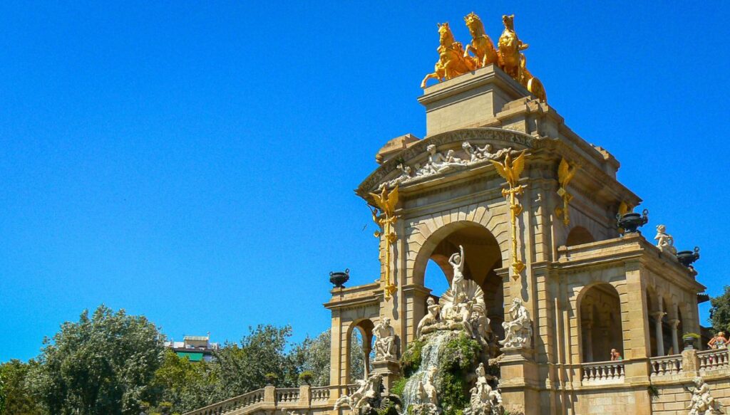 Cascade at Parc de la Ciutadella in Barcelona. There is a bright blue sky and a set of three arches with a set of three gold horses on top. Down below on the ground of the park you see the sculpture at the center of a fountain. 