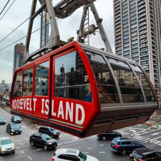 View of the red Roosevelt Island tram car pulling into the station with people standing inside on a cloudy day with the city in back of it.