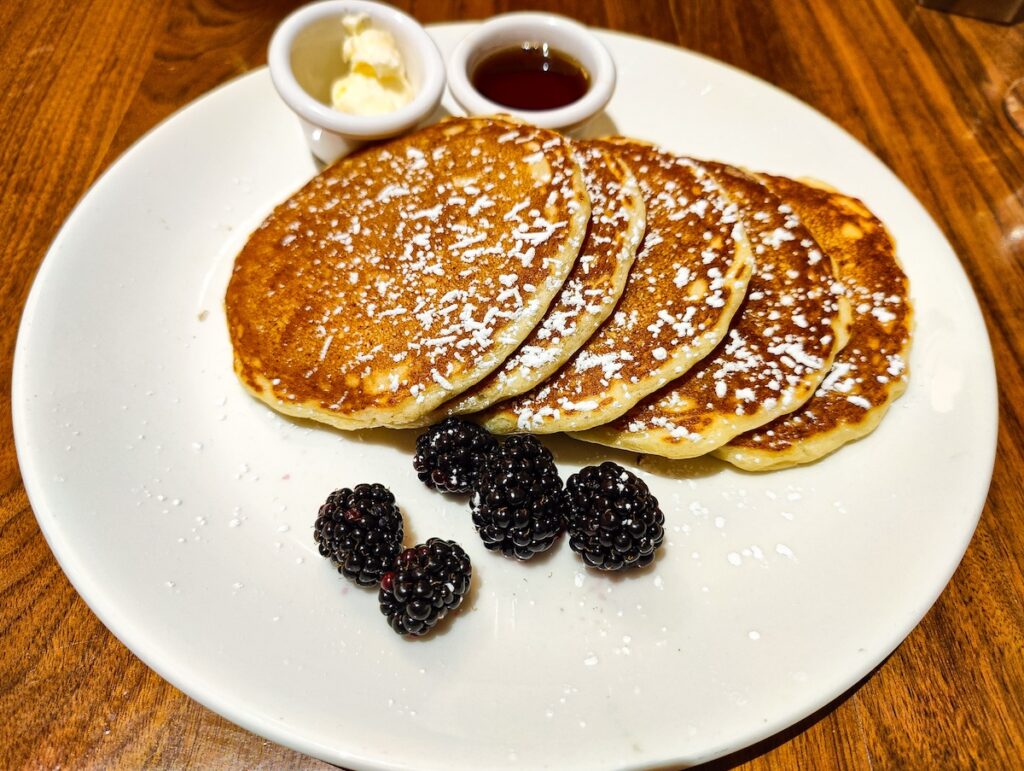 A white plate with five lemon ricotta pancakes with powdered sugar on top. Five black berries sit on the sip and two small containers have maple syrup and butter. The plate sits on a wooden tables. 