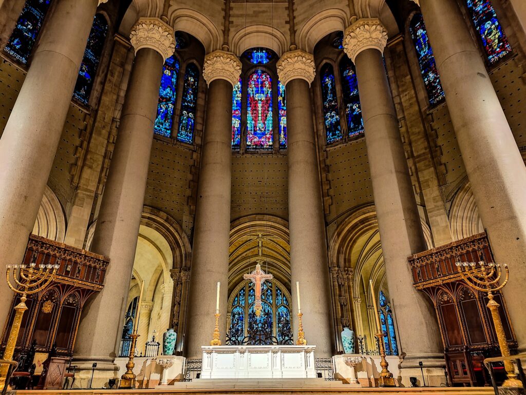 Central altar of St. John the Divine Cathedral. You see stained glass windows at the top and four large stone columns that come down from the ceeiling and that surround the back of the altar. 
