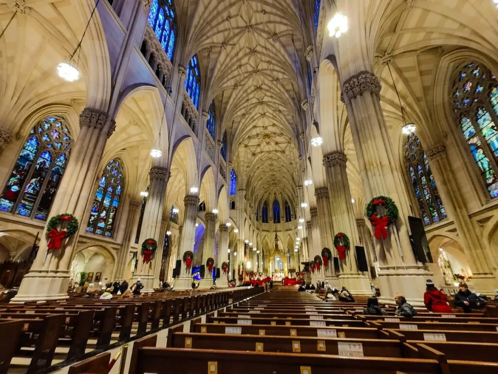 The interior of St. Patrick's Cathedral with wooden pews and columns adorned with green Christmas wreaths with red bows. You can see the vast interior of the church with stained glass windows and the main altar in the distance. 