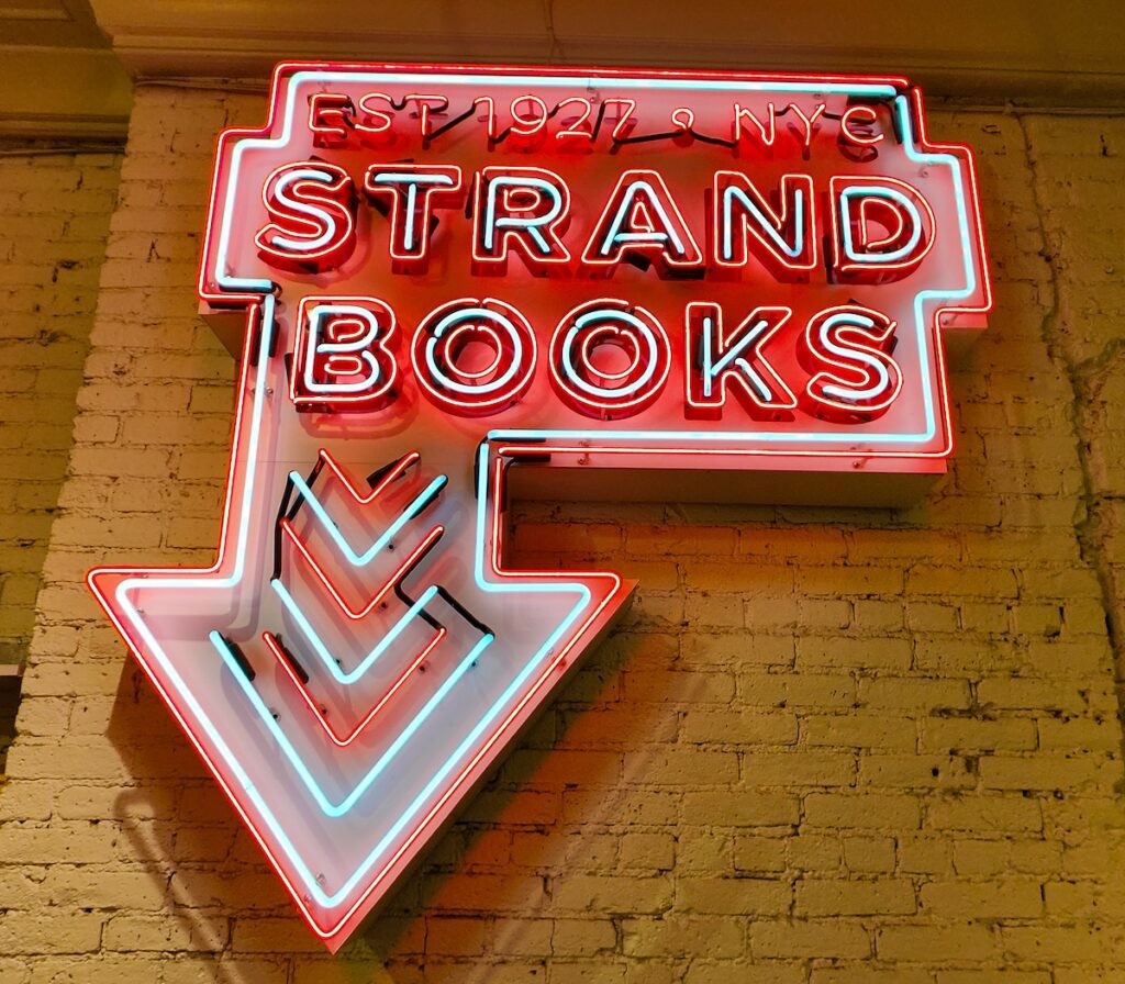 Neon read sign that says Strand books and that it was established in 1927 and there is a red and green arrow pointing down to the stairs at Strand on the Upper West Side. 