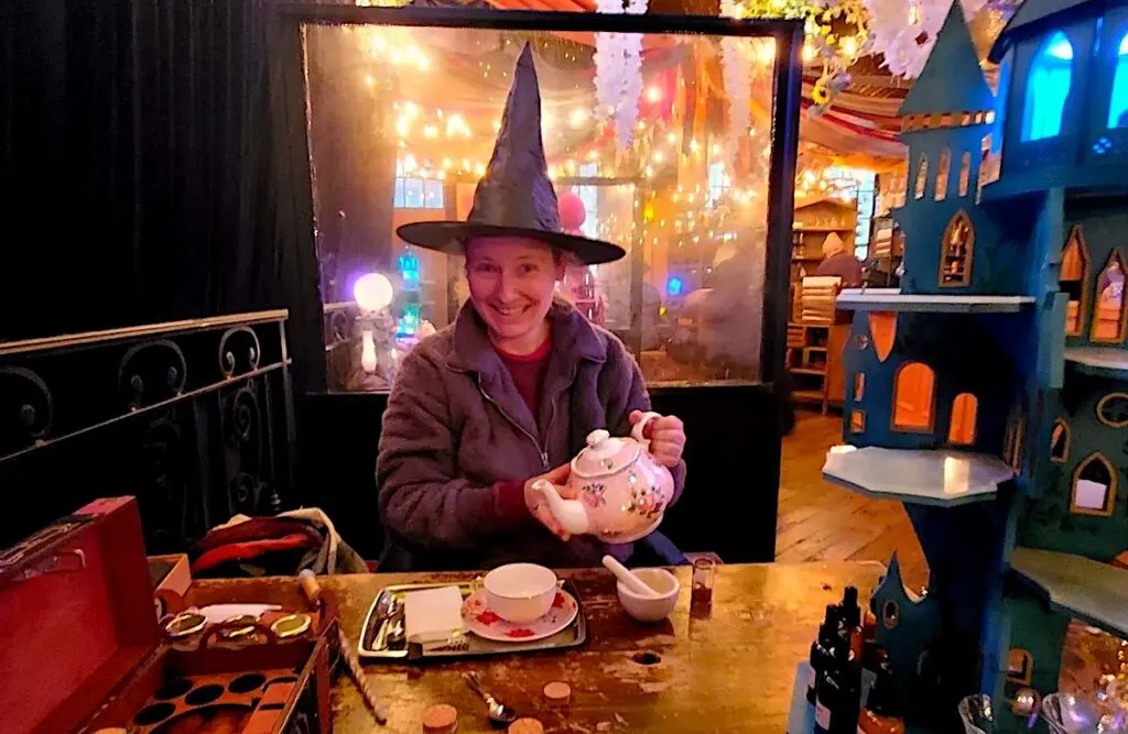 Me wearing a black witch's hat and holding a white pot of tea at the cauldron during afternoon tea.