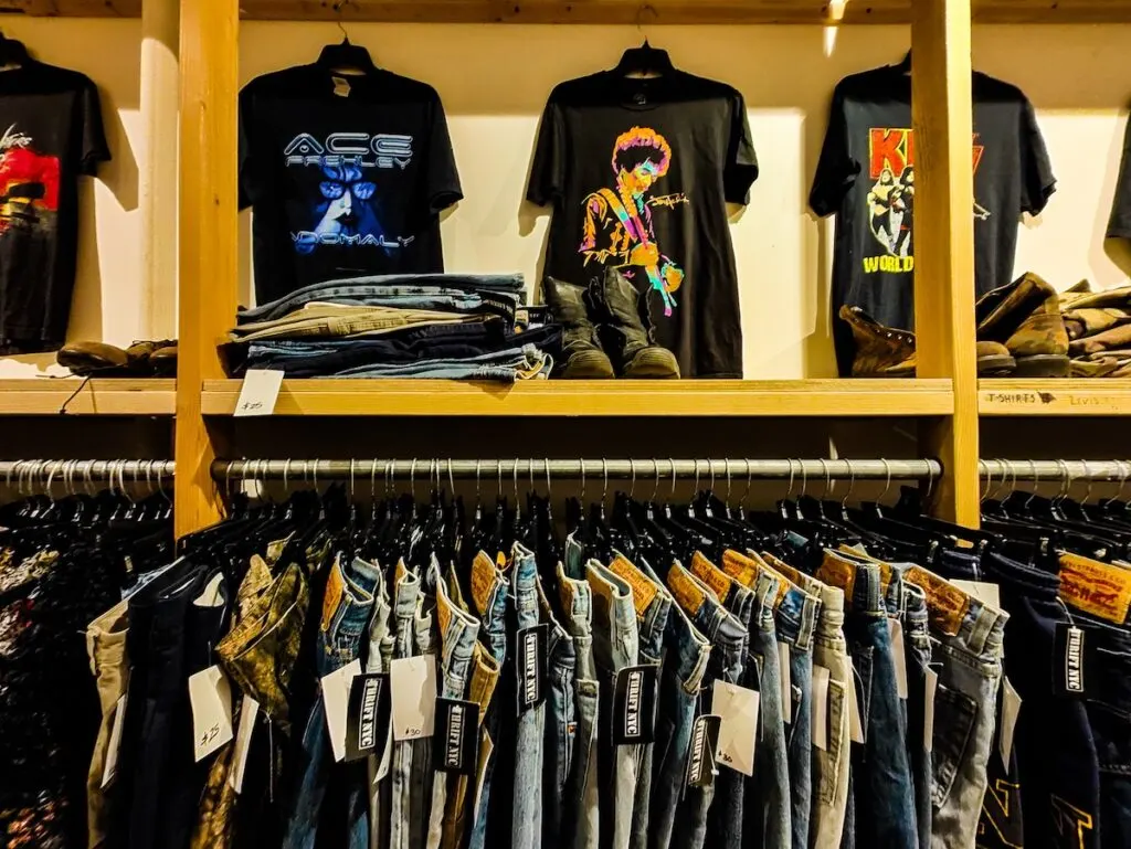 An assortment of back t-shirts on hangers with a whitw background and inside wdooen shelving. Below you see racks of jeans at one of the best thrift stores in NYC. 