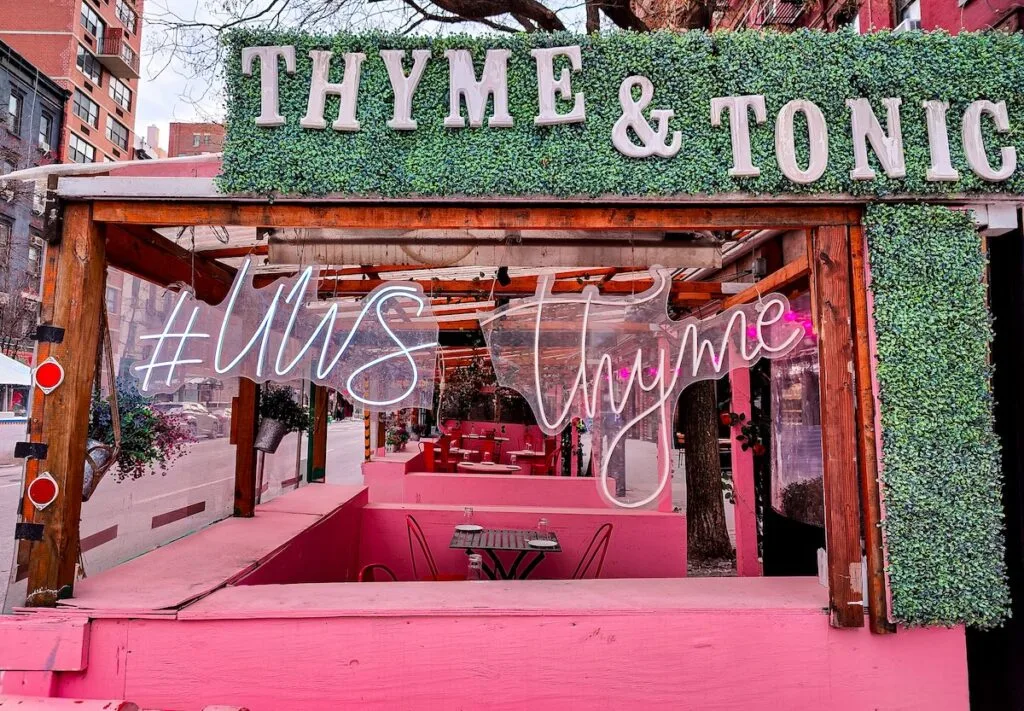 Exterior of Thyme and Tonic. Large white letters with the name of the restaurant sit on an almost moss-like green surface that hangs above an outdoor seating area with vibrant pink walls and wooden beams that connect to the roof.