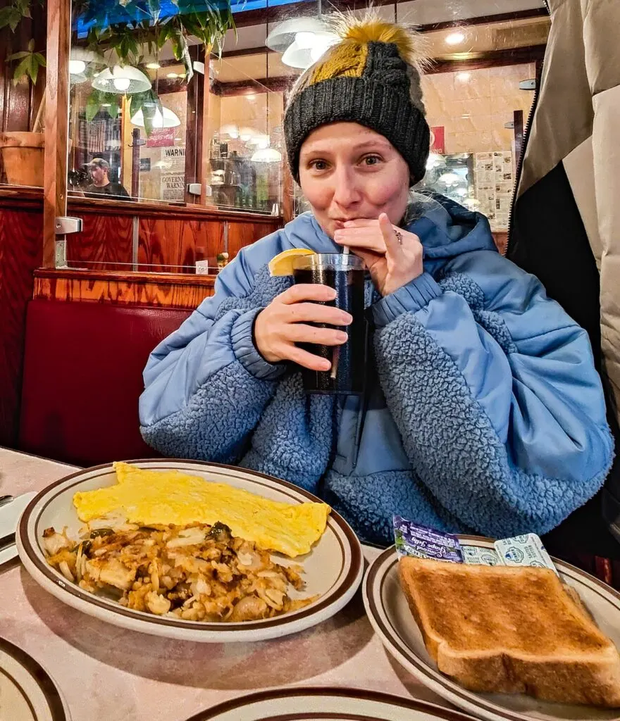 Me sipping on a cup with soda and a lemon. I have a blue fleece and a gray winter hat on. I have an omelet and home fires on a plate in front of me and wheat toast on the table next to me. I ma sitting in a red booth with a tan coat hanging next to me. 