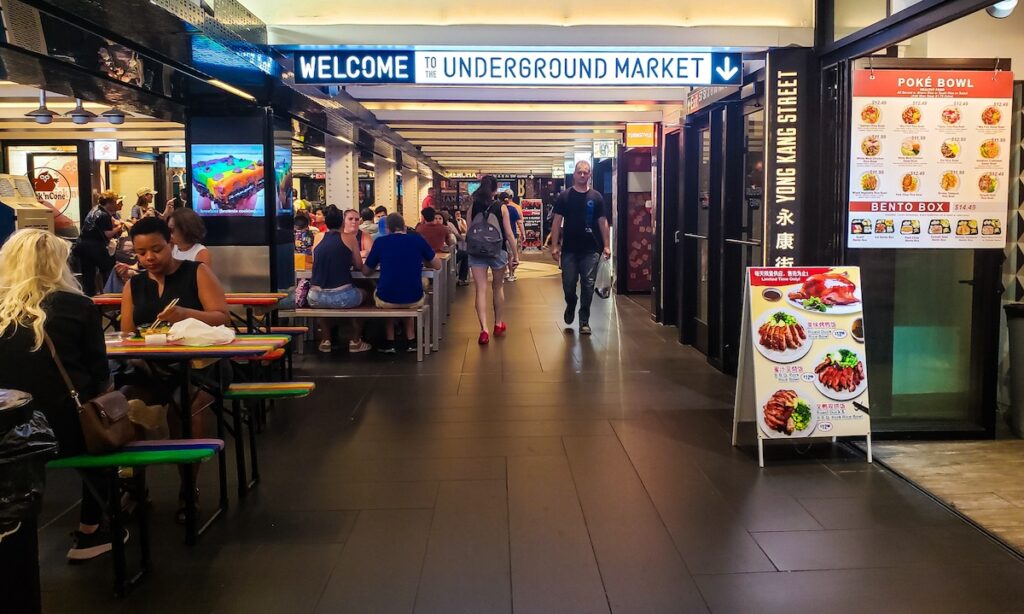 People walking beneath a white underground market sign at Turnstyle. There are food stalls on either side with food signs on the right and tables with people sitting on the left. . 