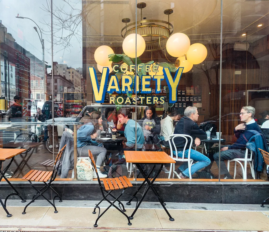 A view through the exterior window of Variety Coffee Roasters. People are sitting inside and bubble lights hang from the ceiling with the name of the shop printed on the window in blue and gold. 