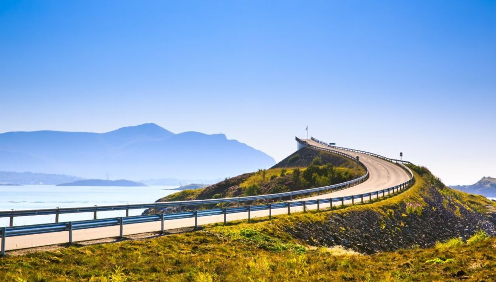A guardrail sits around a curving road in Norway with green grass around it and a road that climbs to stunning views. 