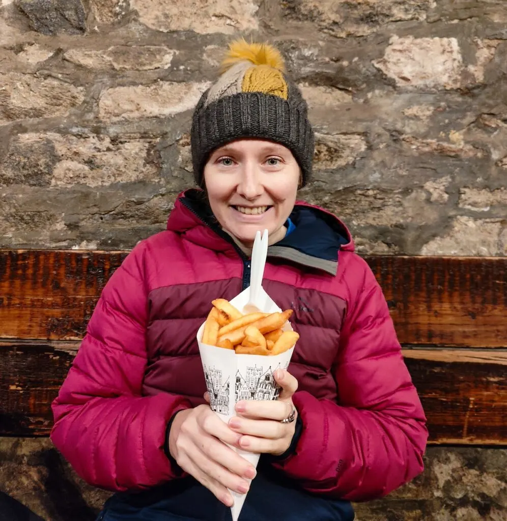 Me holding a a cone of Belgian fires with a white, plastic fork in it. I have a gray and yellow winter hat on and am wearing a pink jacket with a stone wall and wooden railing behind me. So, is Antwerp worth visiting? Well, for the fries it is.
